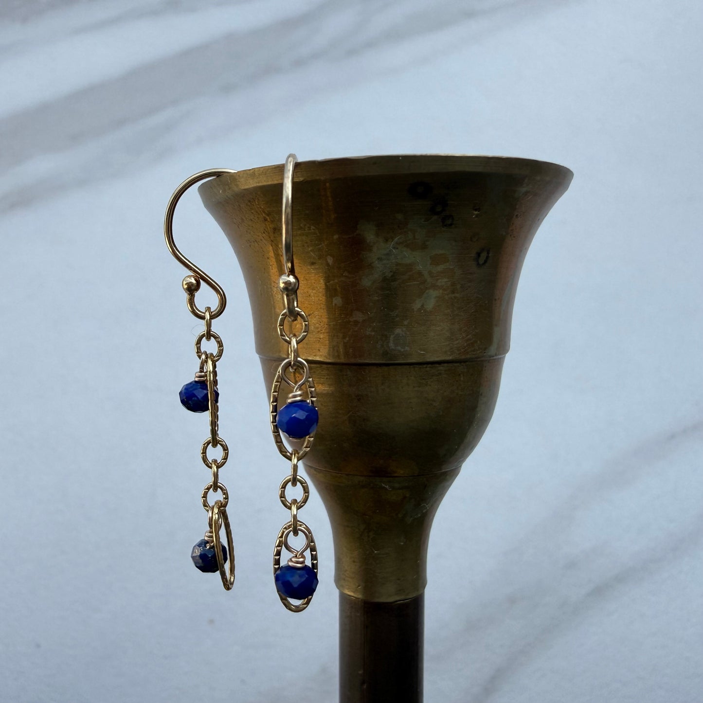 A pair of gold-toned dangle earrings with deep blue bead accents, hanging from a brass candle holder against a light gray marble background.