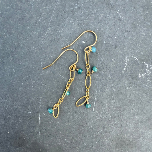 A pair of gold dangle earrings with small turquoise blue beads, displayed on a dark gray textured surface.
