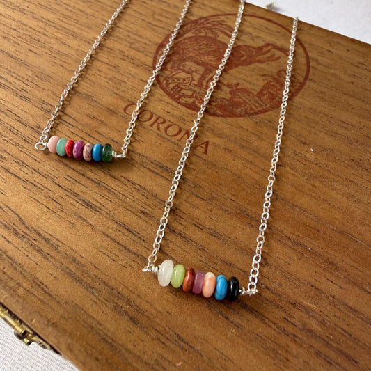 Two delicate silver chain necklaces with small horizontal rows of colorful round beads, displayed on a wooden box with a Corona logo.