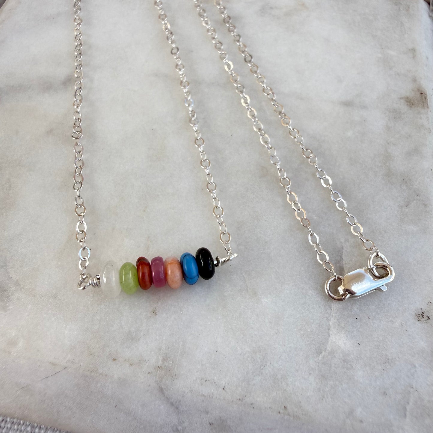 A silver chain necklace with a horizontal bar of seven colorful round beads in green, red, pink, orange, blue, black, and white, displayed on a light marble surface.