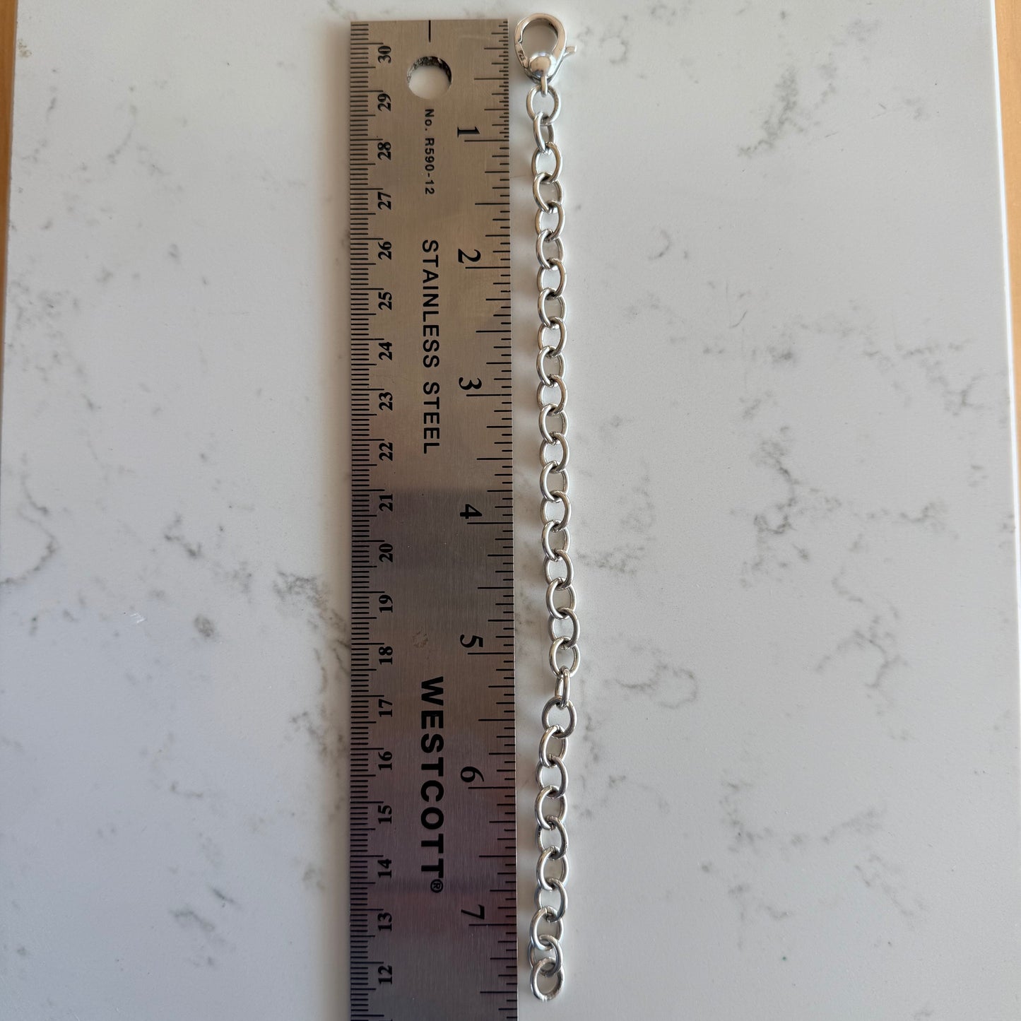 A silver metal link chain extension with a small ring at one end, shown vertically next to a stainless steel ruler on a white marble-patterned surface.