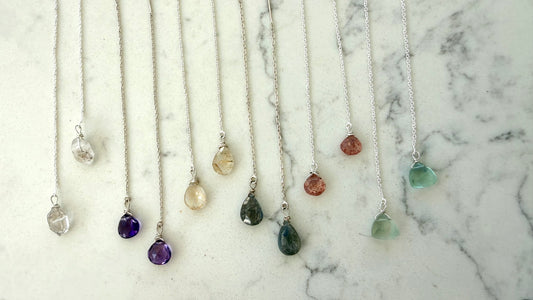 Nine silver pendant necklaces with teardrop-shaped gemstone pendants are arranged in a row on a white marble surface. The necklaces feature various colored stones including clear, purple, yellow, green, brown, and light blue.