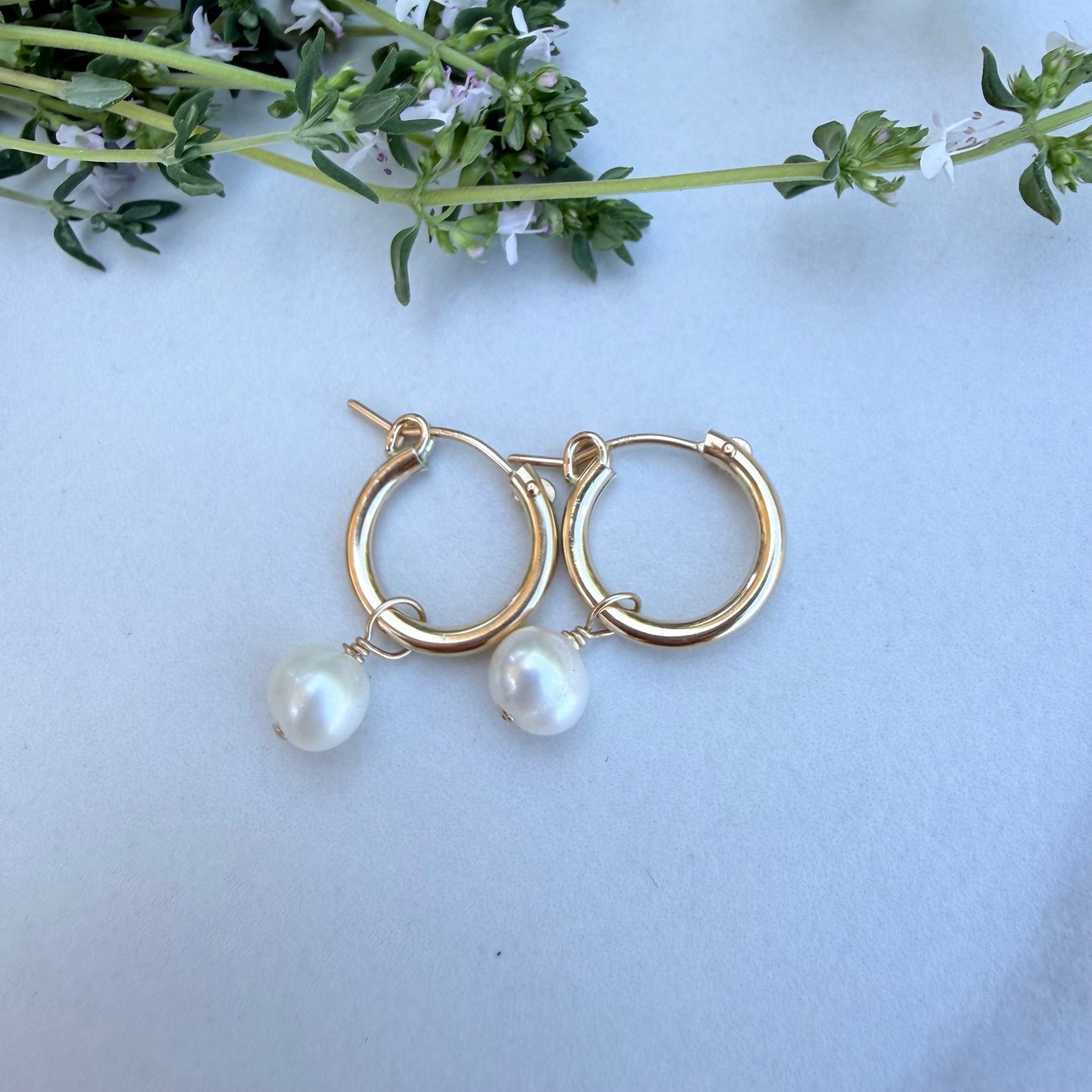 Gold hoop earrings with pearl drops on a light blue background with greenery.