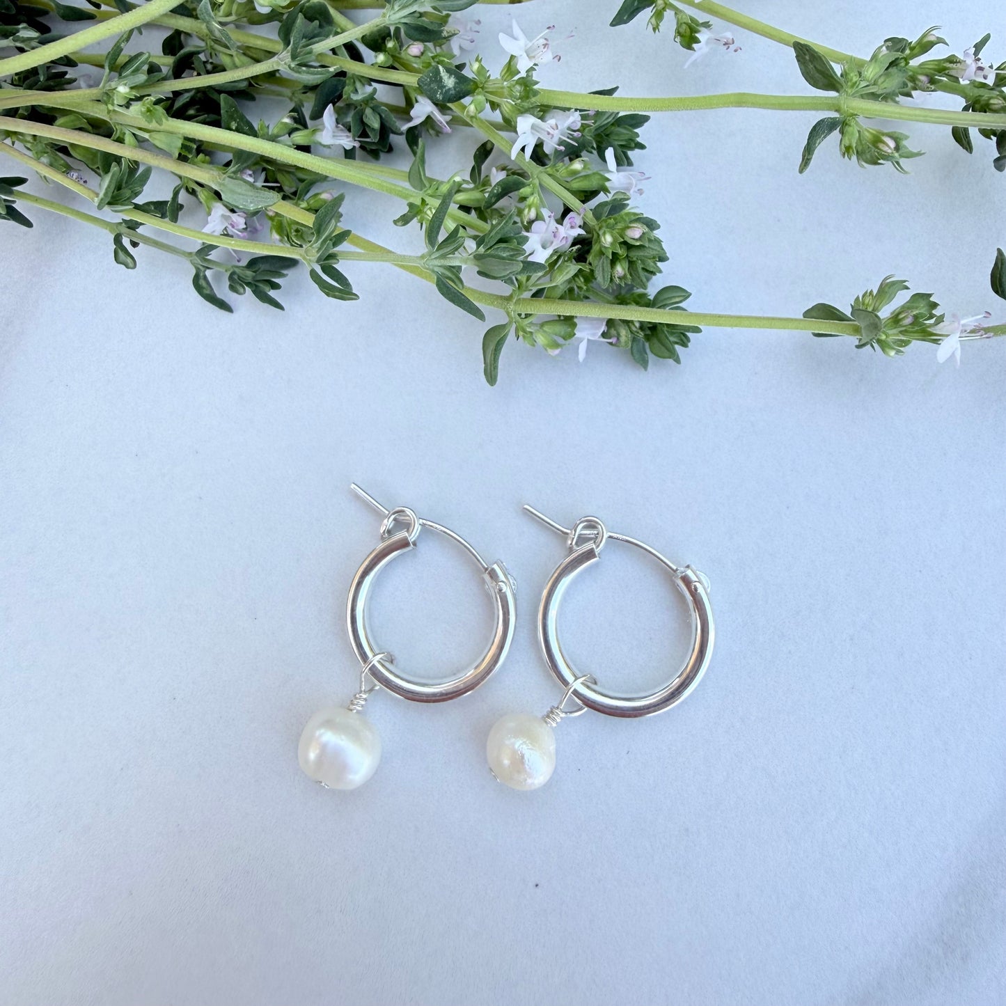 Silver hoop earrings with pearl drops on a light blue background with greenery.