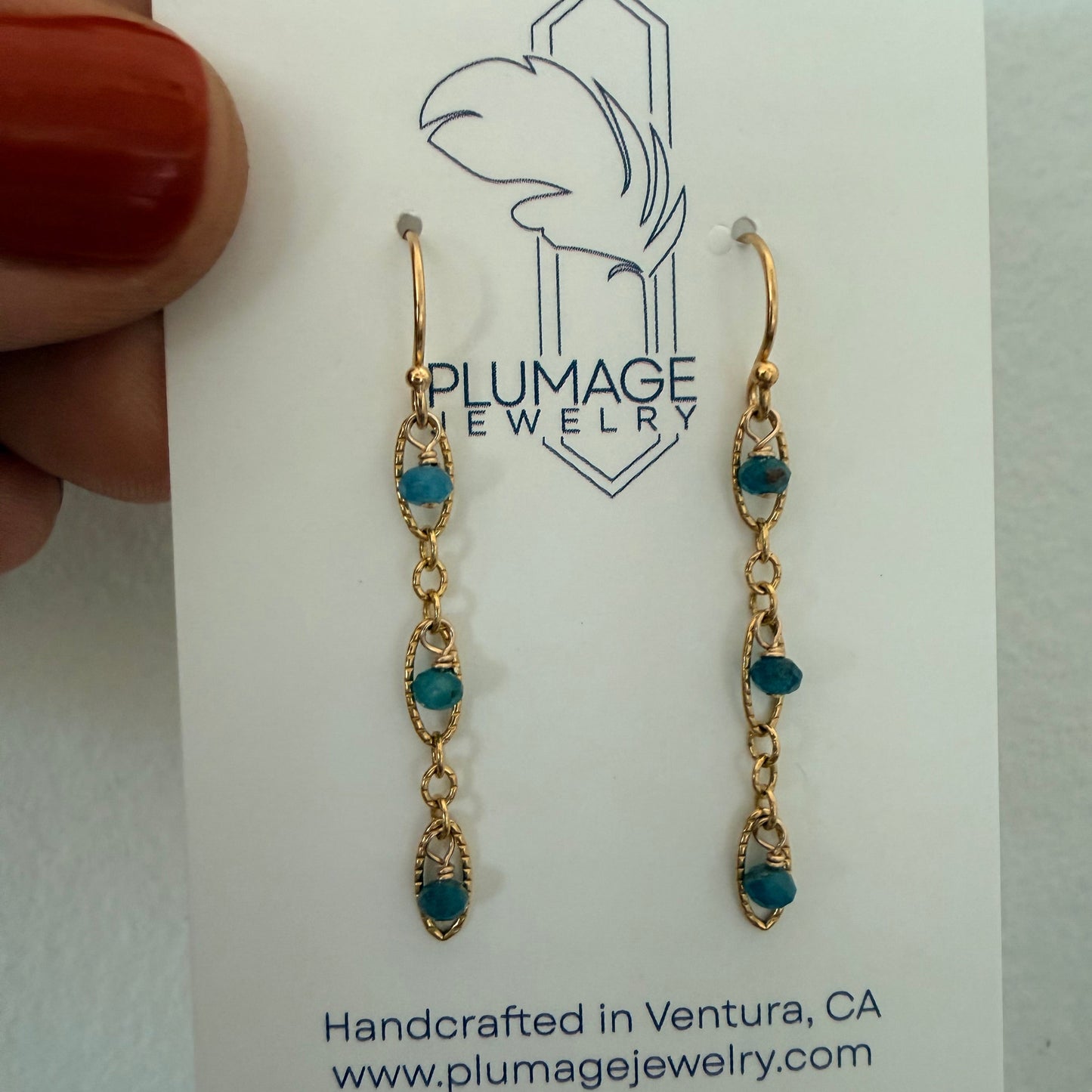 A pair of gold drop earrings with small teal blue beads, displayed on a white card from Plumage Jewelry. A hand with red nail polish holds the card on the left side.