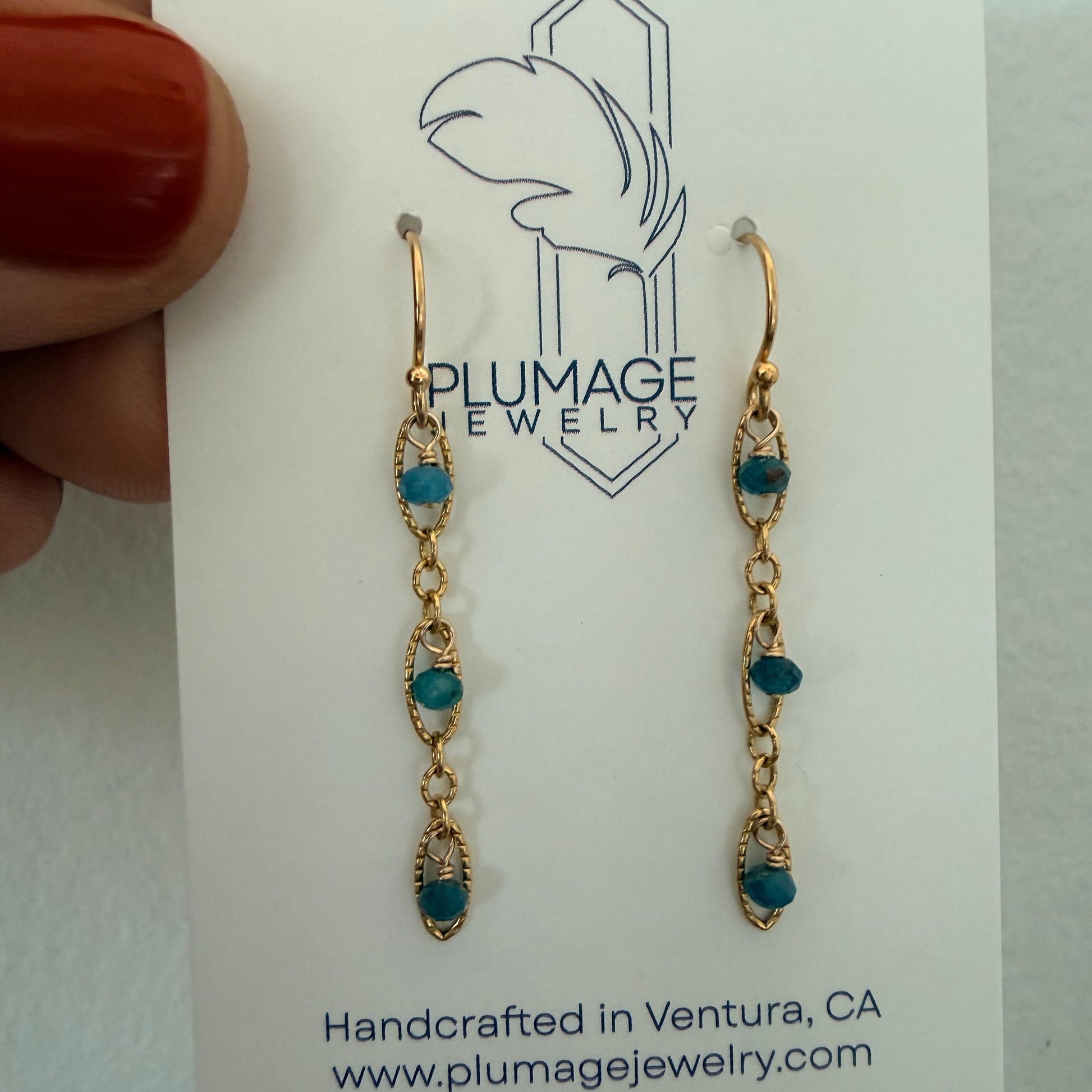 A pair of gold drop earrings with small teal blue beads, displayed on a white card from Plumage Jewelry. A hand with red nail polish holds the card on the left side.