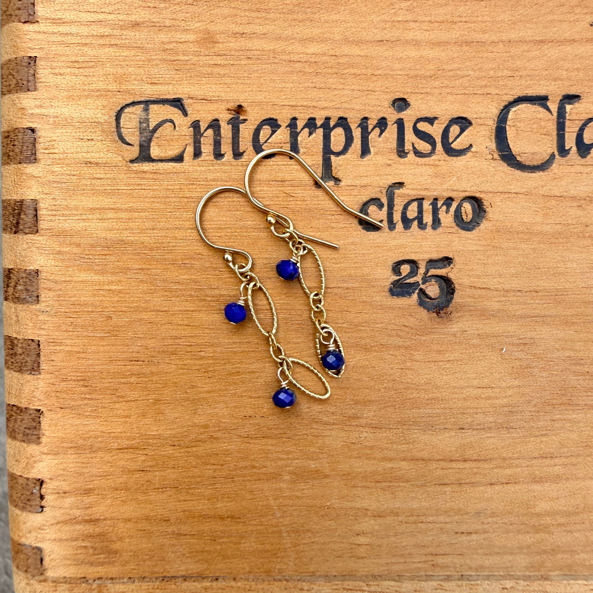 A pair of handmade dangle earrings with gold-toned wirework and deep blue beads, displayed on a wooden box engraved with 'Enterprise Claro 25'.