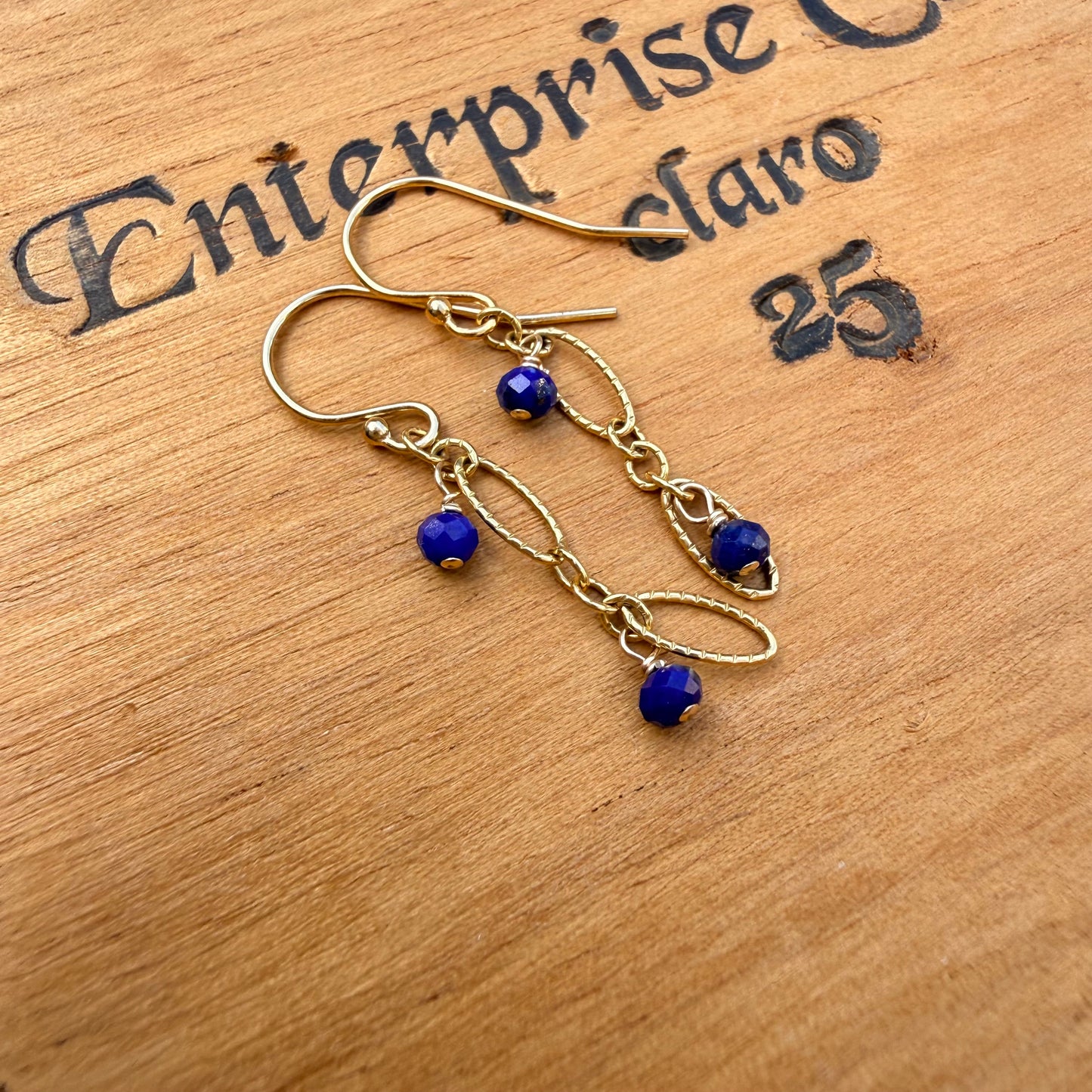 A pair of gold-toned dangle earrings with small blue faceted beads, displayed on a wooden surface with black printed text in the background.
