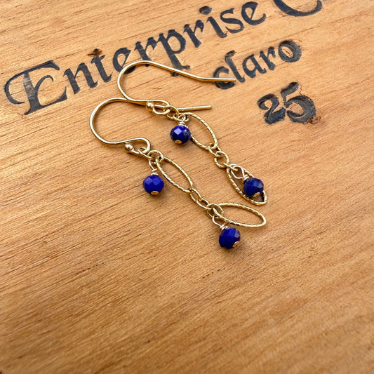A pair of gold-toned dangle earrings with small blue faceted beads, displayed on a wooden surface with black printed text in the background.