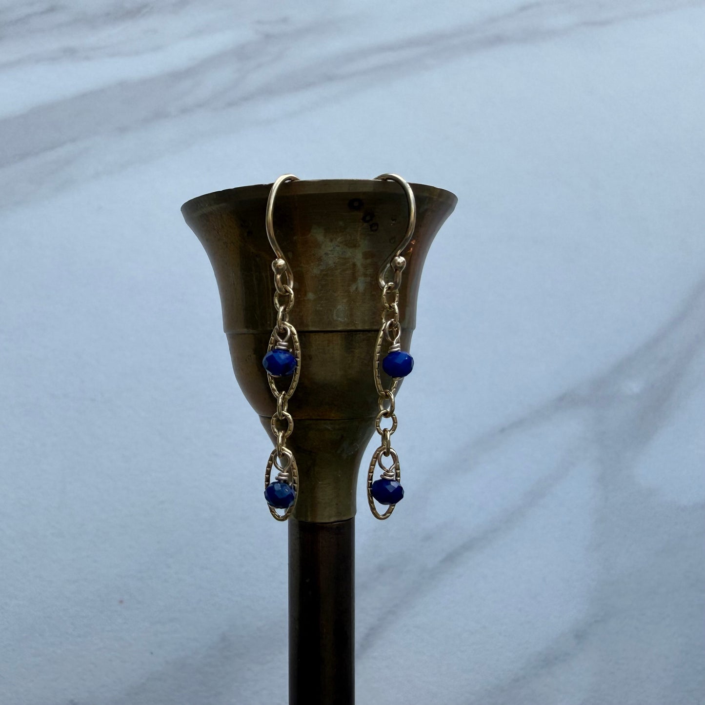 Gold-toned dangle earrings with blue beads and clear rhinestone accents, hanging from a brass candle holder against a light gray marble background.