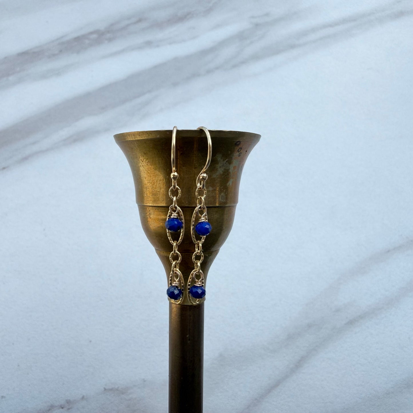 A pair of gold-toned dangle earrings with small blue beads, hanging from a brass candle holder against a light gray marble background.