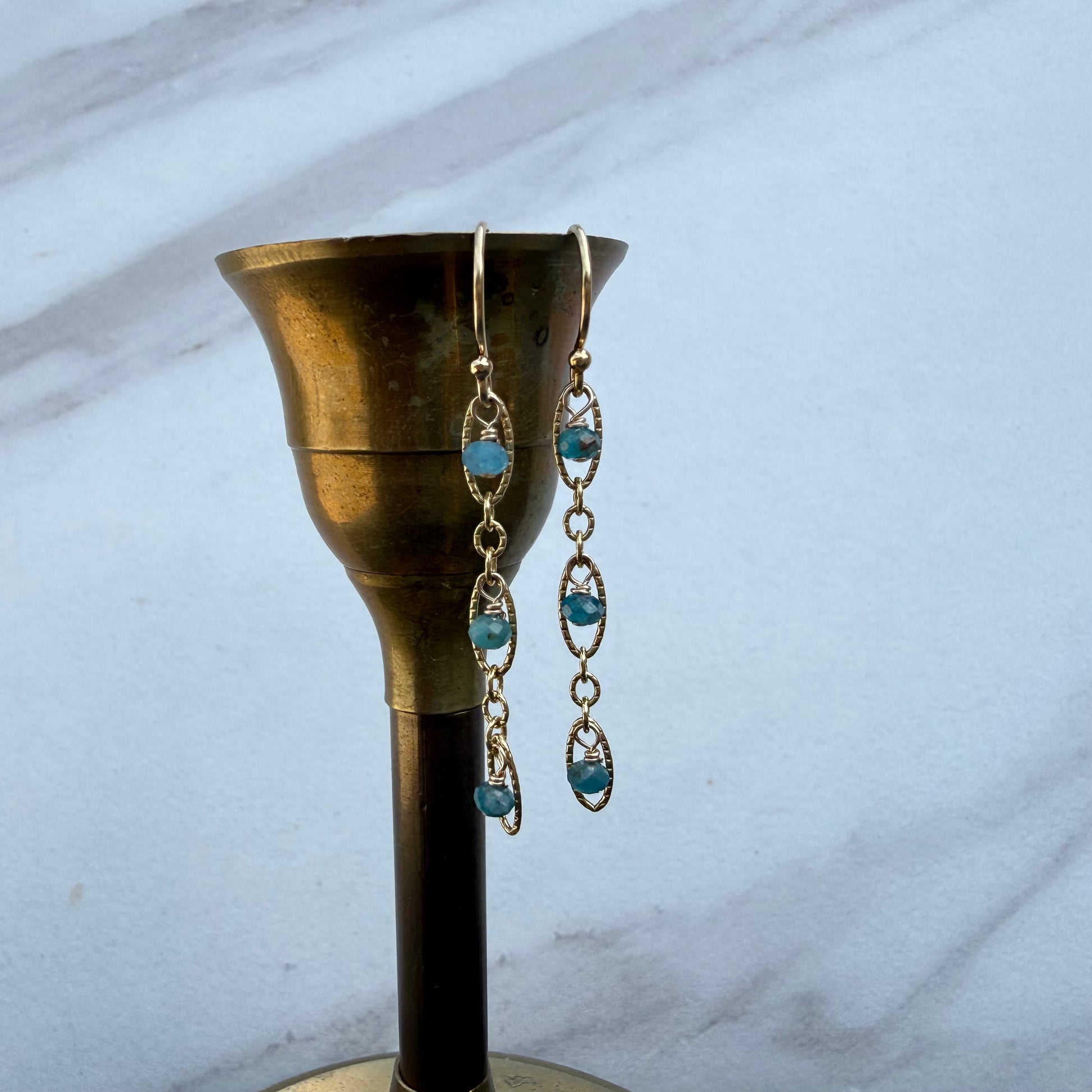 A pair of long dangling earrings with light blue oval gemstones and gold-toned chain links, hanging from a brass candle holder against a light marble background.