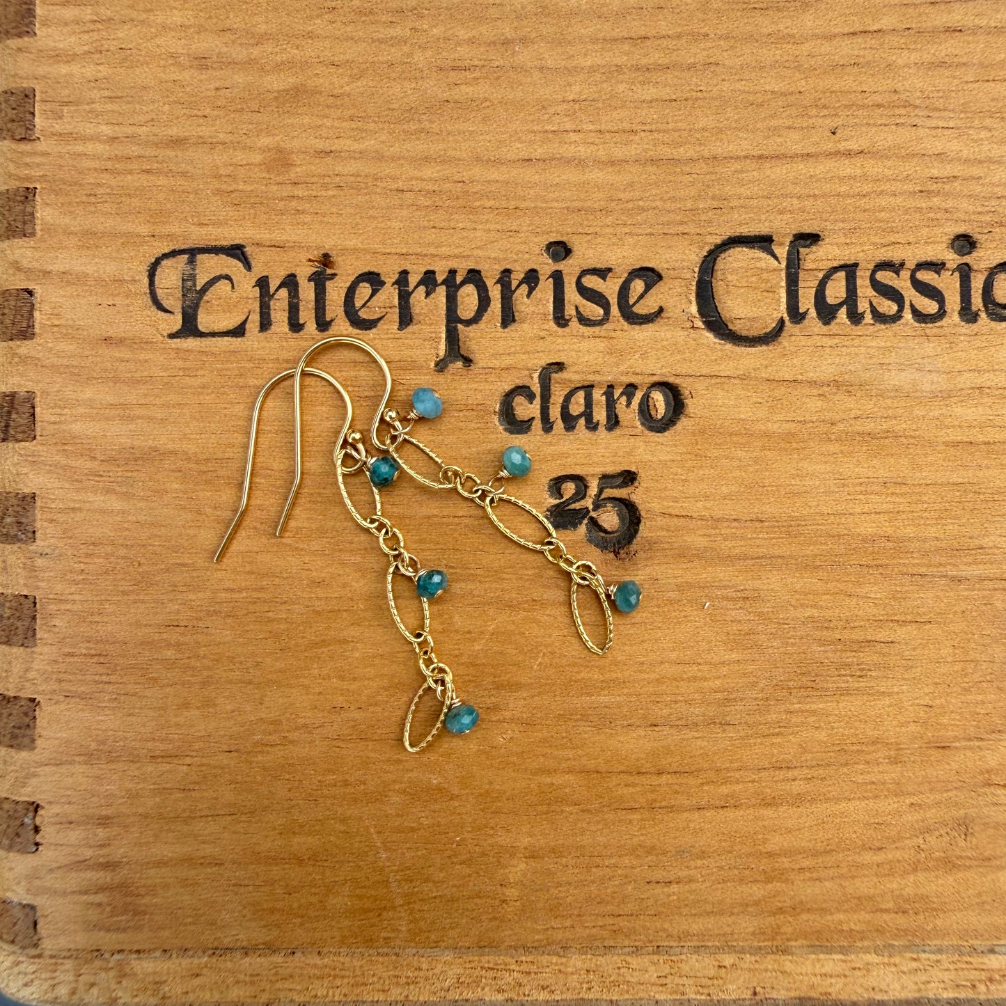 A pair of gold-toned dangle earrings with small turquoise-blue beads, displayed on a wooden box engraved with the text 'Enterprise Classic claro 25'.