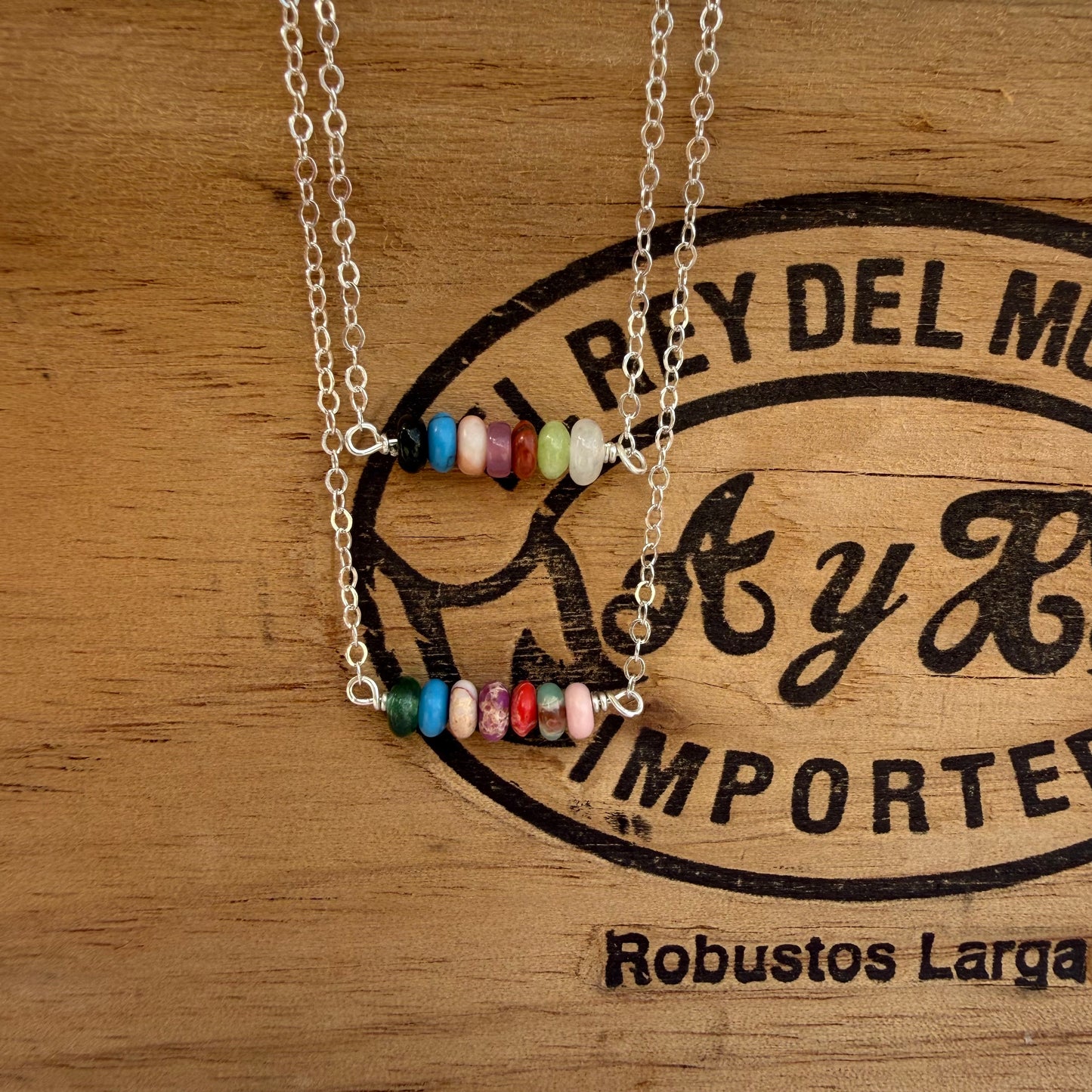 Two silver chain necklaces with small colorful bead bars, displayed on a wooden surface with a black printed logo.