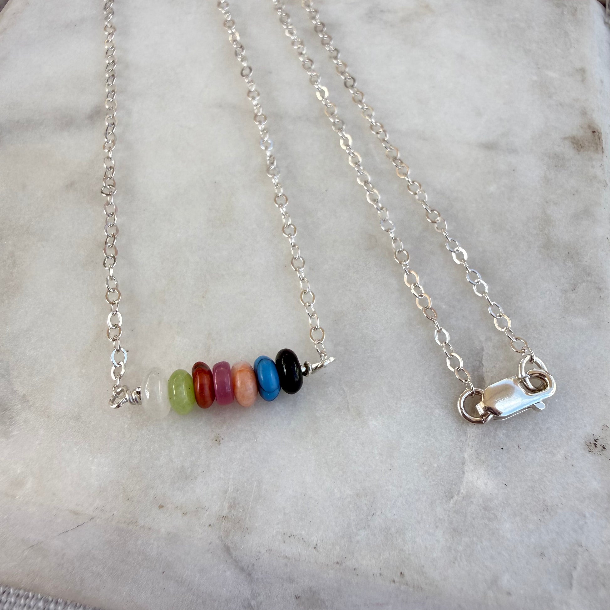 A silver chain necklace with a horizontal bar of seven colorful round beads in green, red, pink, orange, blue, black, and white, displayed on a light marble surface.