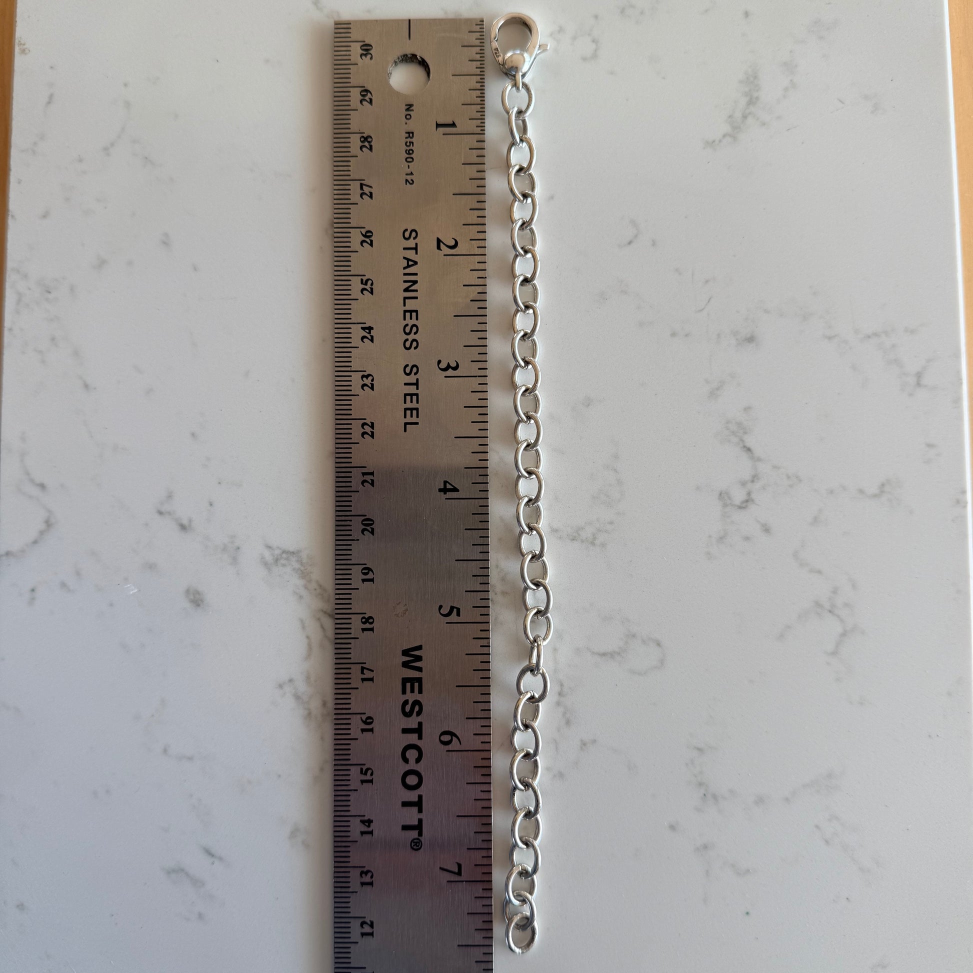 A silver metal link chain extension with a small ring at one end, shown vertically next to a stainless steel ruler on a white marble-patterned surface.